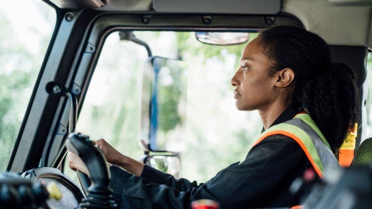 Driver in a safety vest seated in a Mack truck cab, looking ahead through a large side window.
